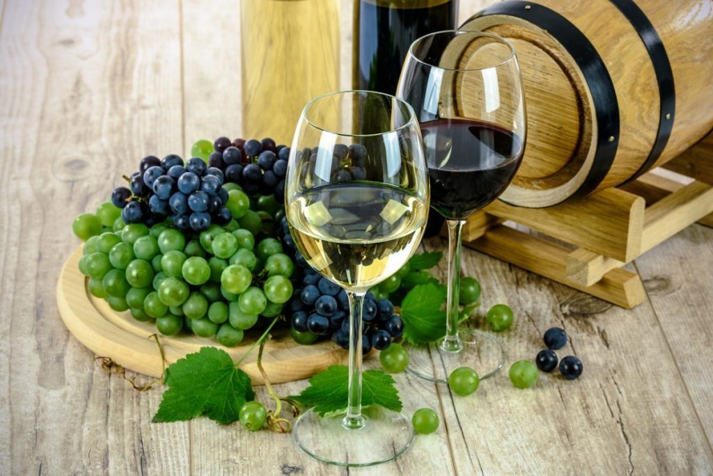 Taça de vinho branco e tinto com uvas verdes e roxas, barril de madeira e garrafas ao fundo, em mesa de madeira