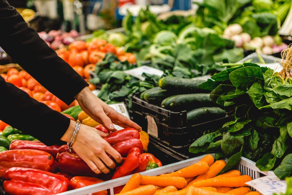 Mãos a escolher pimentos vermelhos e cenouras frescas no mercado, com espinafres e abóboras ao fundo — escolha