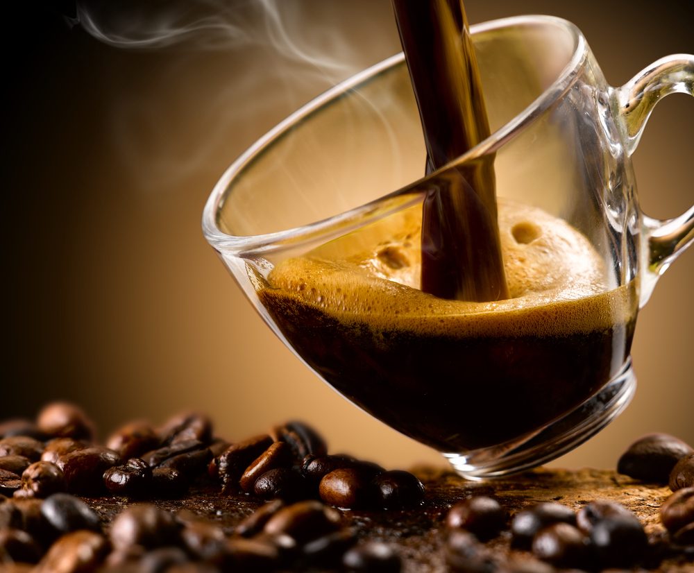 Café preto a ser vertido em chávena de vidro, com grãos torrados à volta — bebida estimulante que pode afetar o sono