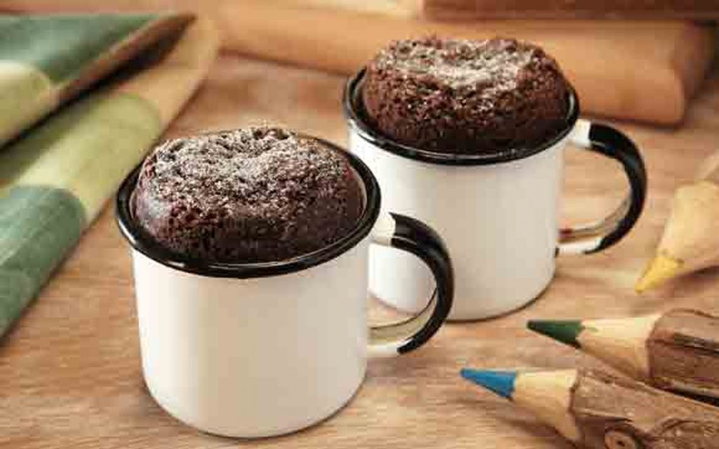 Dois mini bolos de chocolate em canecas brancas com borda preta, sobre mesa de madeira, ao lado de lápis de cor