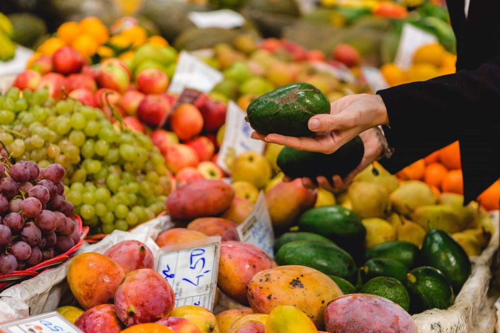 Mão a escolher abacates frescos num mercado de frutas variadas, incluindo uvas, maçãs e mangas, em contexto