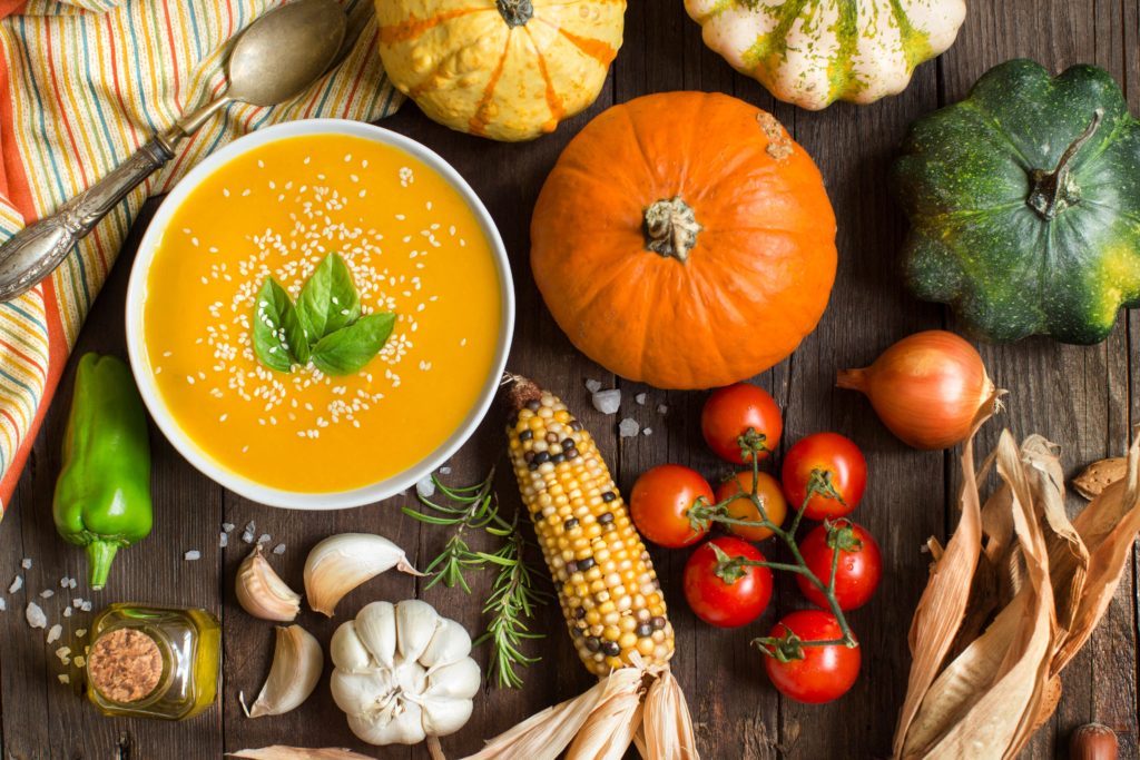 Sopa de abóbora com sementes e manjericão, acompanhada de tomate, alho, milho e pimenta verde, dispostos sobre mesa