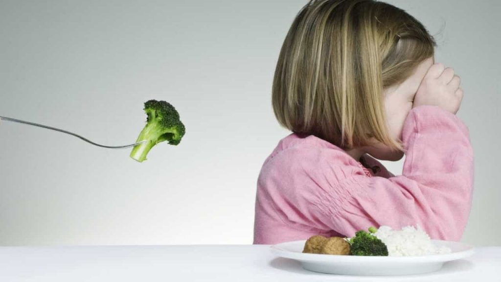 Criança com seletividade alimentar recusa brócolos, prato com arroz e almôndegas à frente, gesto de rejeição